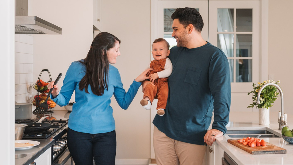 Dad holding child and mum cooking dinner in kitchen